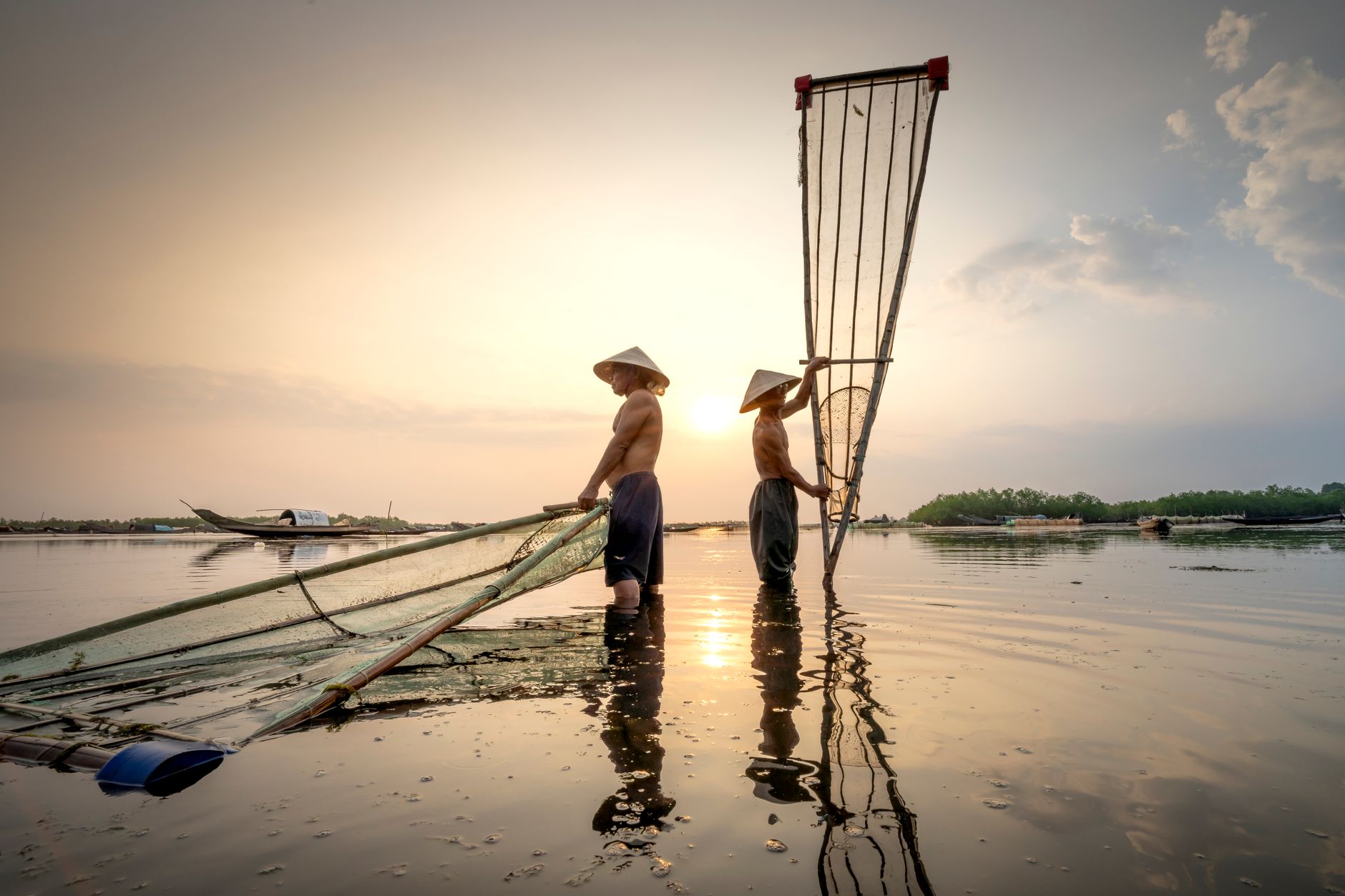 Asian fishermen in Vietnamese hats with fishing nets standing in river under cloudy sky at sunset