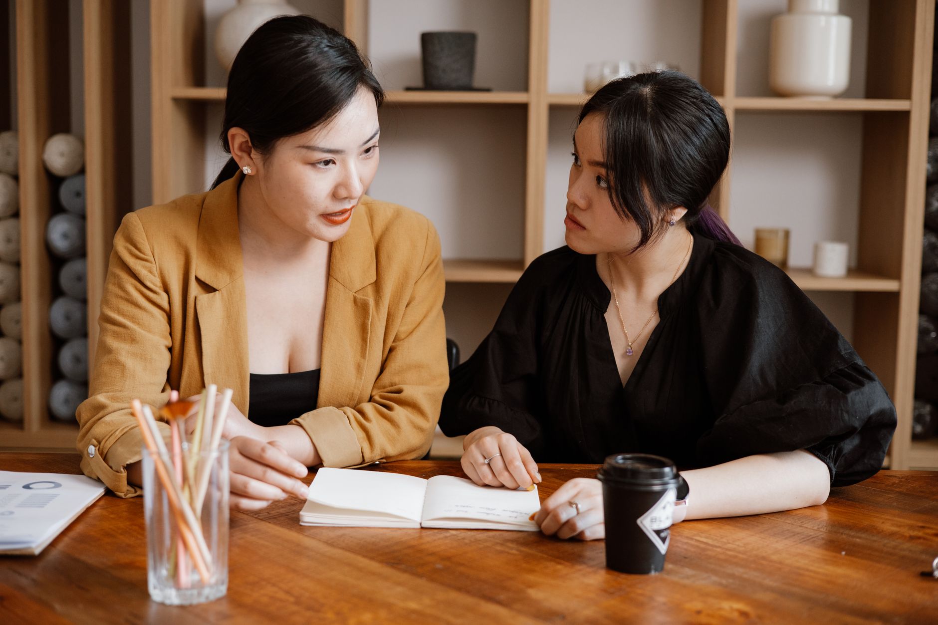 Two women engaged in a business meeting at an office desk with notebooks and coffee.