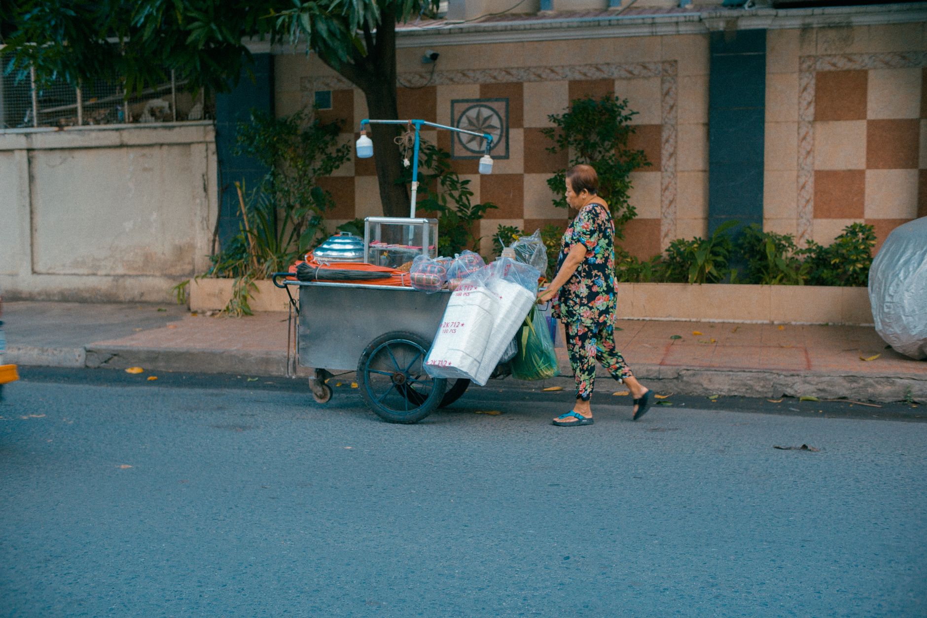 An elderly woman street vendor pushing a cart on a Phnom Penh street.