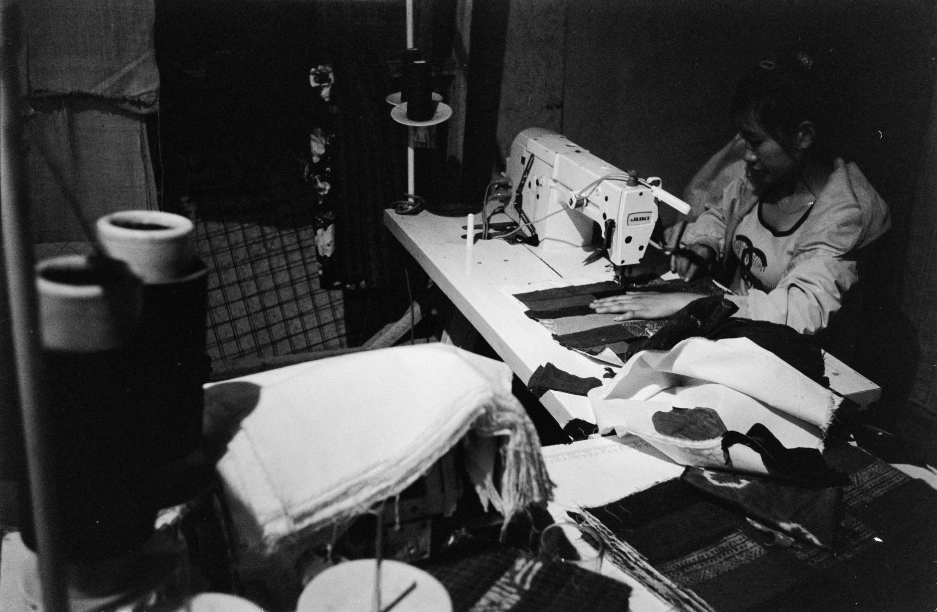 Photo en noir et blanc d’une personne cousant du tissu à une machine à coudre dans un atelier sombre, avec des tissus et des bobines au premier plan.