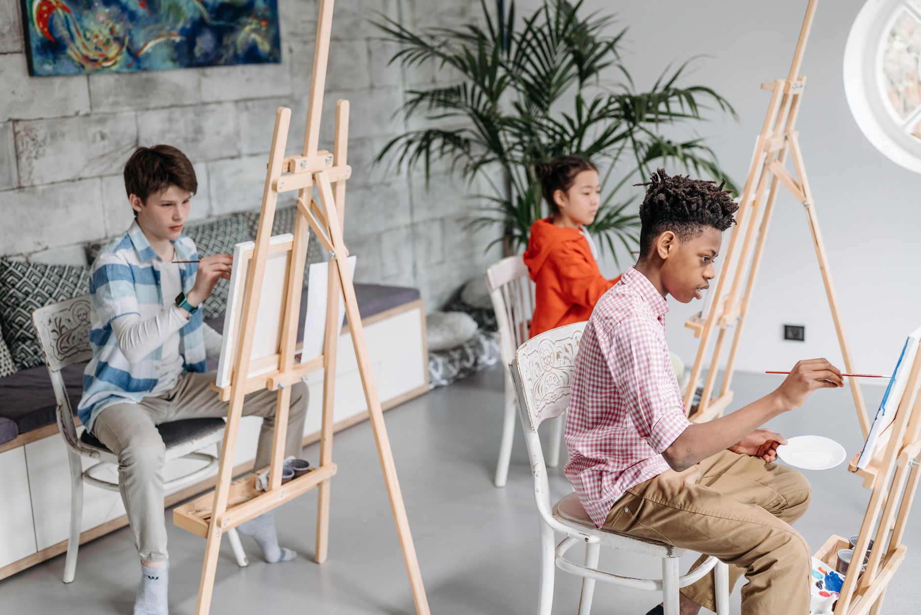 Trois jeunes peignent devant des chevalets en bois dans un atelier lumineux, avec une banquette, une grande plante et un tableau au mur.
